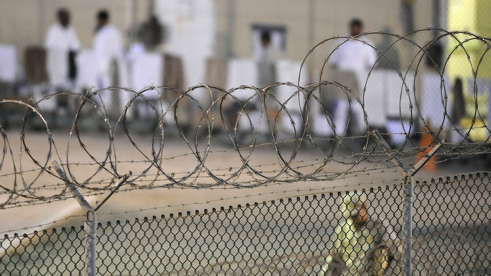 A close-up shot of a barbed-wire fence; a guard walks behind it, out of focus.