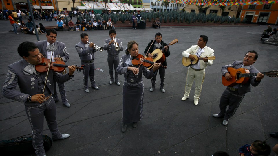 A mariachi band stands in a semi circle playing guitars, violins, and trumpets. A woman sands in the middle playing the violin.