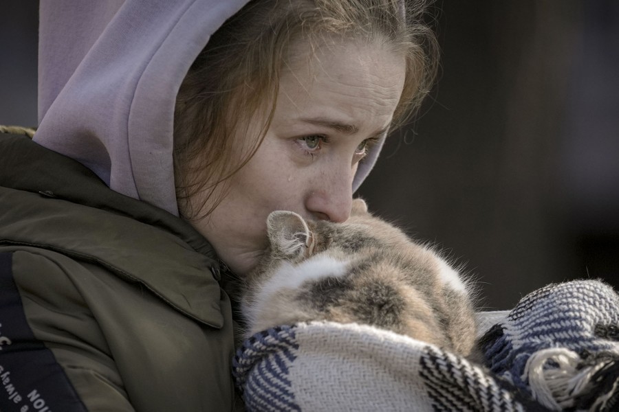 A woman leans down to kiss the head of a cat she carries, with a teardrop visible on her cheek.