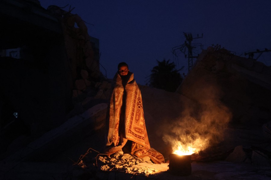 A Palestinian boy wrapped in a blanket keeps warm next to a fire atop rubble.