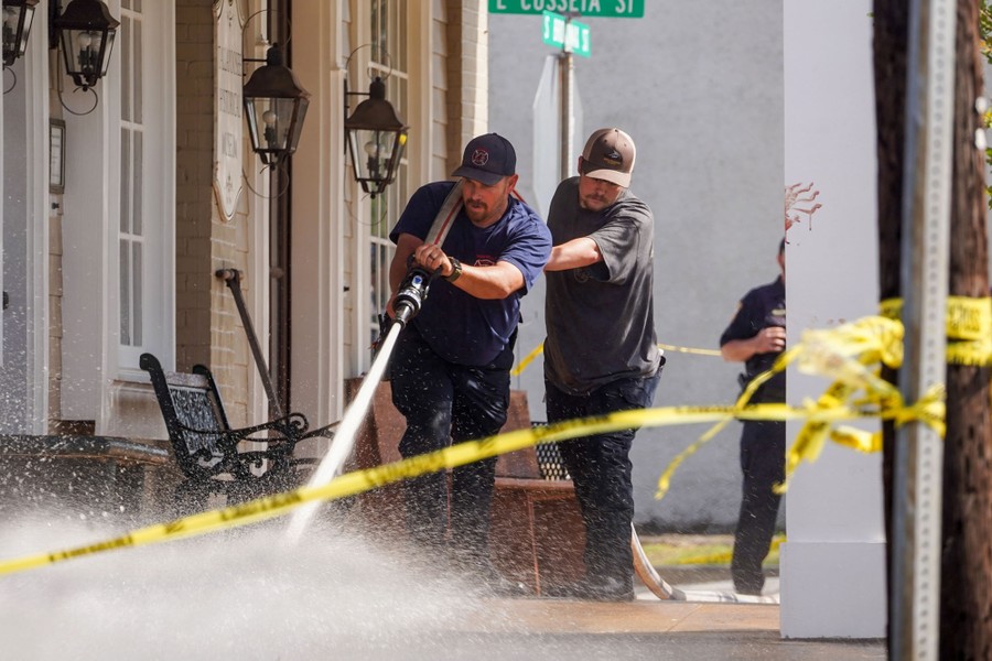 Two firefighters use a large hose to spray water on a sidewalk.