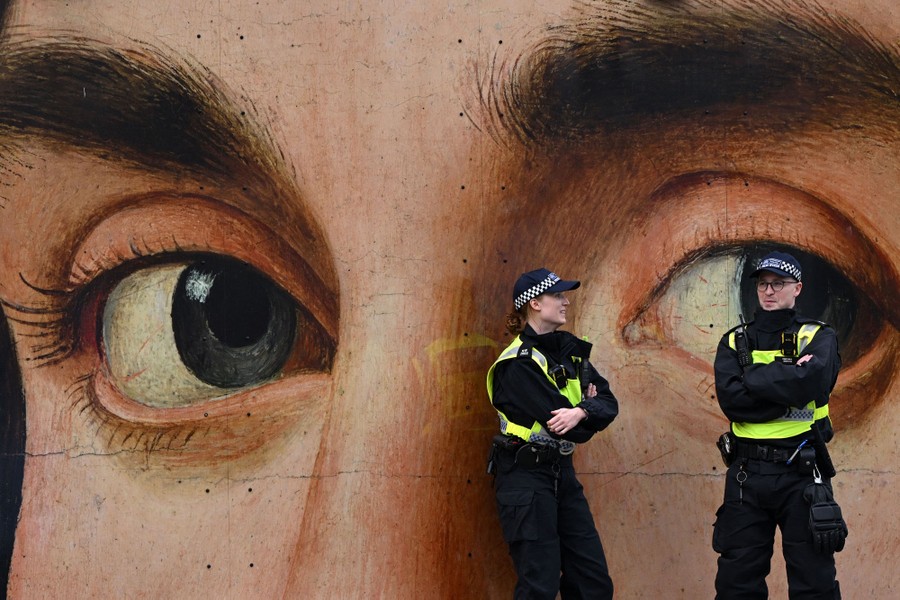 Two police officers stand in front of a large painting of a man's eyes.