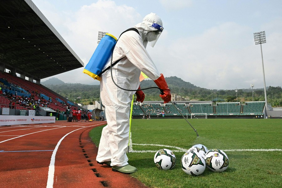 A person wearing protective gear sprays disinfectant on several soccer balls.