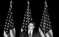Black-and-white photo of Ron DeSantis speaking into a microphone, in front of three American flags