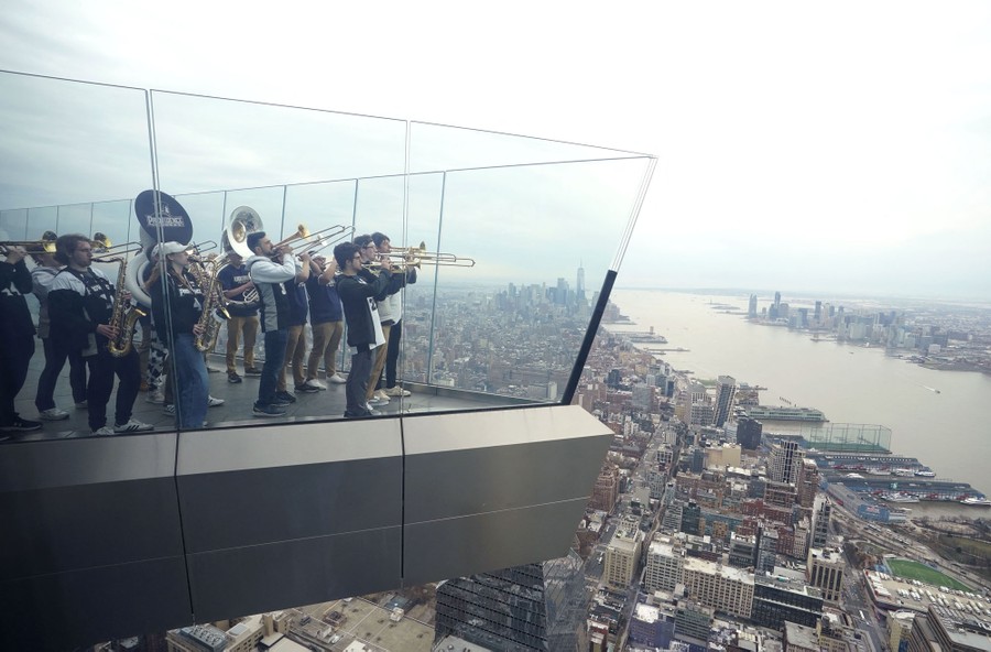 Members of a marching band play their instruments while standing at the edge of a high observation deck above New York City.