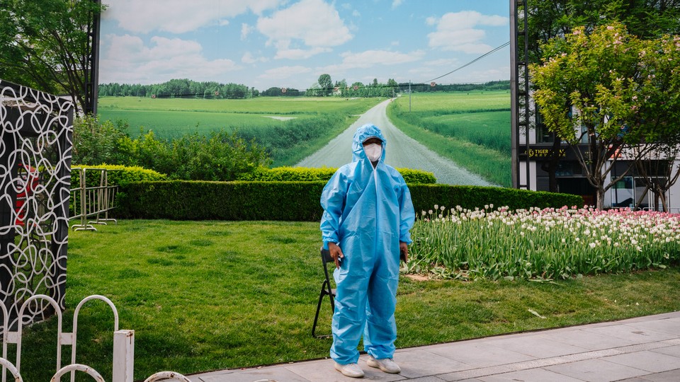 A security guard wearing a Hazmat suit stands in front of a patch of green and a large photo of rolling fields in Beijing