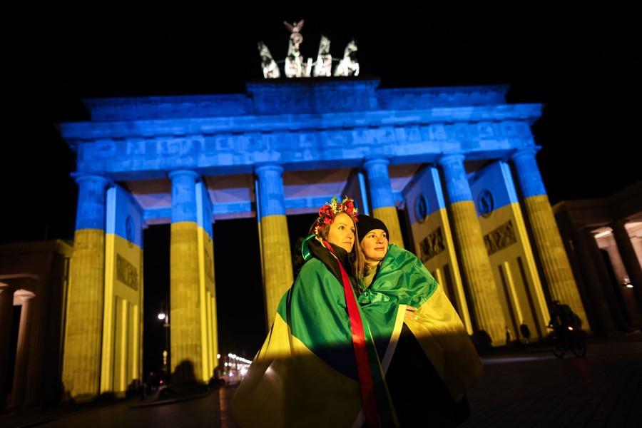 Two women draped in flags pose for a photo in front of an illuminated monument.