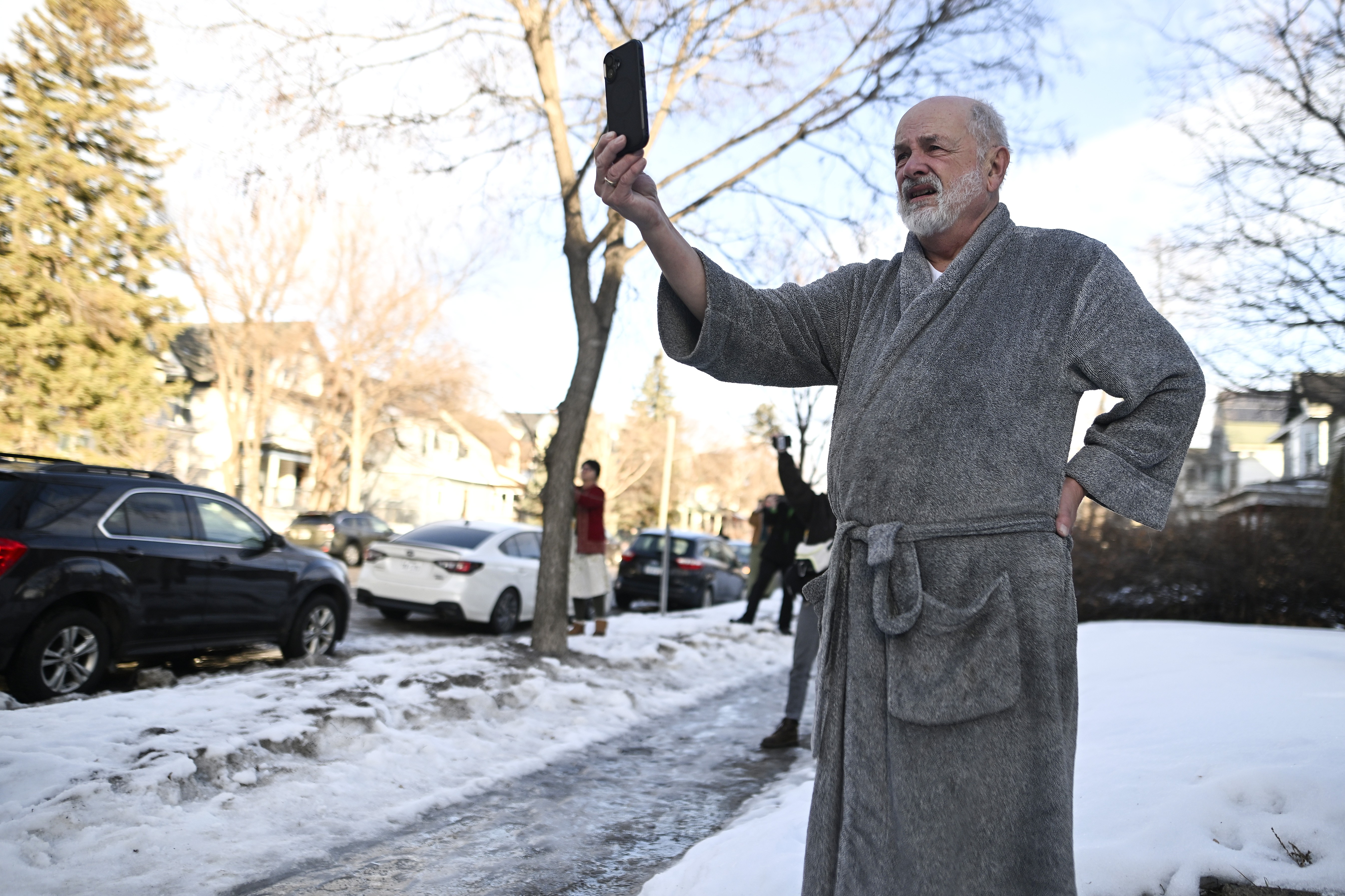 A man wearing a bathrobe stands in a snow-covered front yard, holding up a phone to take video of an immigration enforcement action.