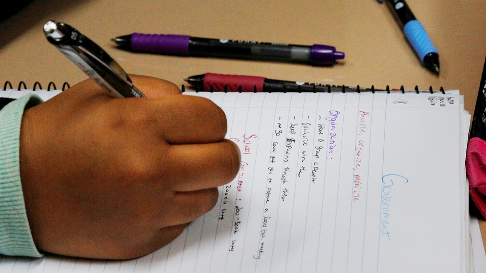 A student holds a pencil above a sheet of notebook paper. The notes appear to be about the government