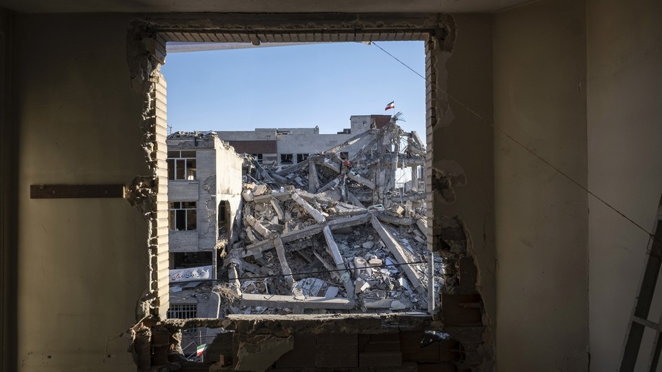 A photograph through a jagged hole in a wall shows a collapsed building across the street with an Iranian flag flying atop it.
