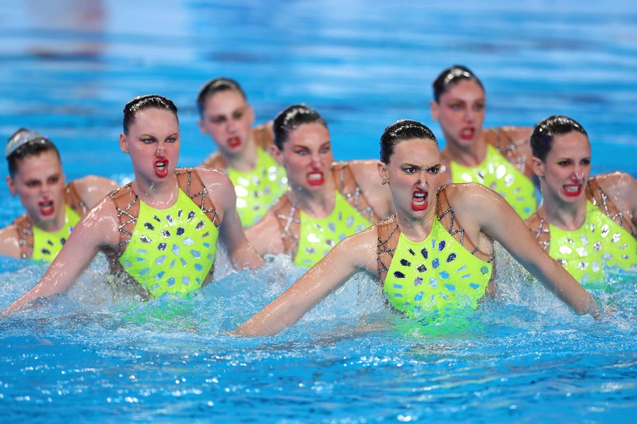 A team of synchronized swimmers makes intense faces while performing.