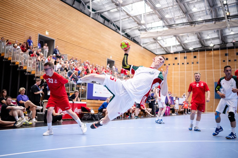 An athlete jumps to throw a handball as others stand nearby.