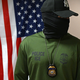 An image of a person wearing a shirt that reads "Police ICE" and who has their face fully covered with a neck gaiter and head covered with a Yankees baseball cap, standing in front of an American flag.