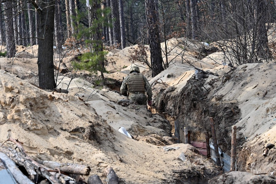 A soldier walks through a newly dug trench in a forest.