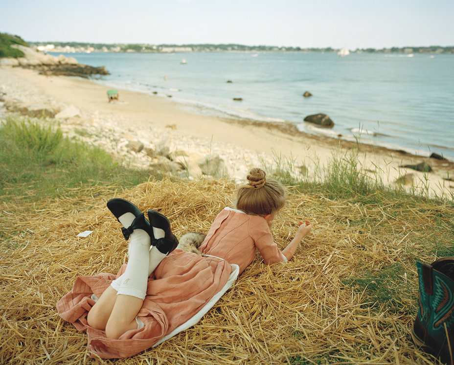 Photo of a young woman in pink dress lying on stomach, with legs crossed wearing long white socks and black shoes, in pile of straw looking out at the beach and water at Gloucester, MA 