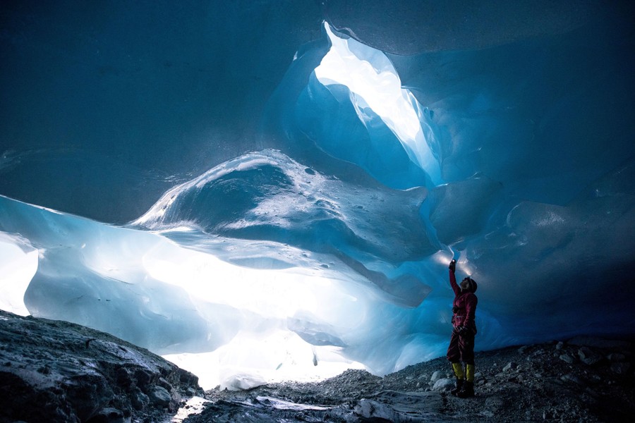 A person stands in a large cave formed by glacial ice above and hard rock below.