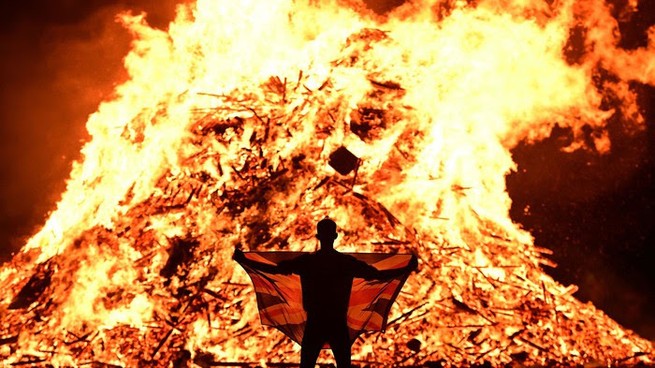 A person stands in front of a large bonfire. 