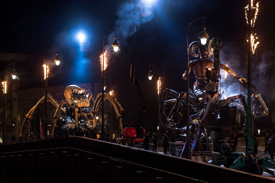 Two giant mechanical puppets meet on a city bridge at night.