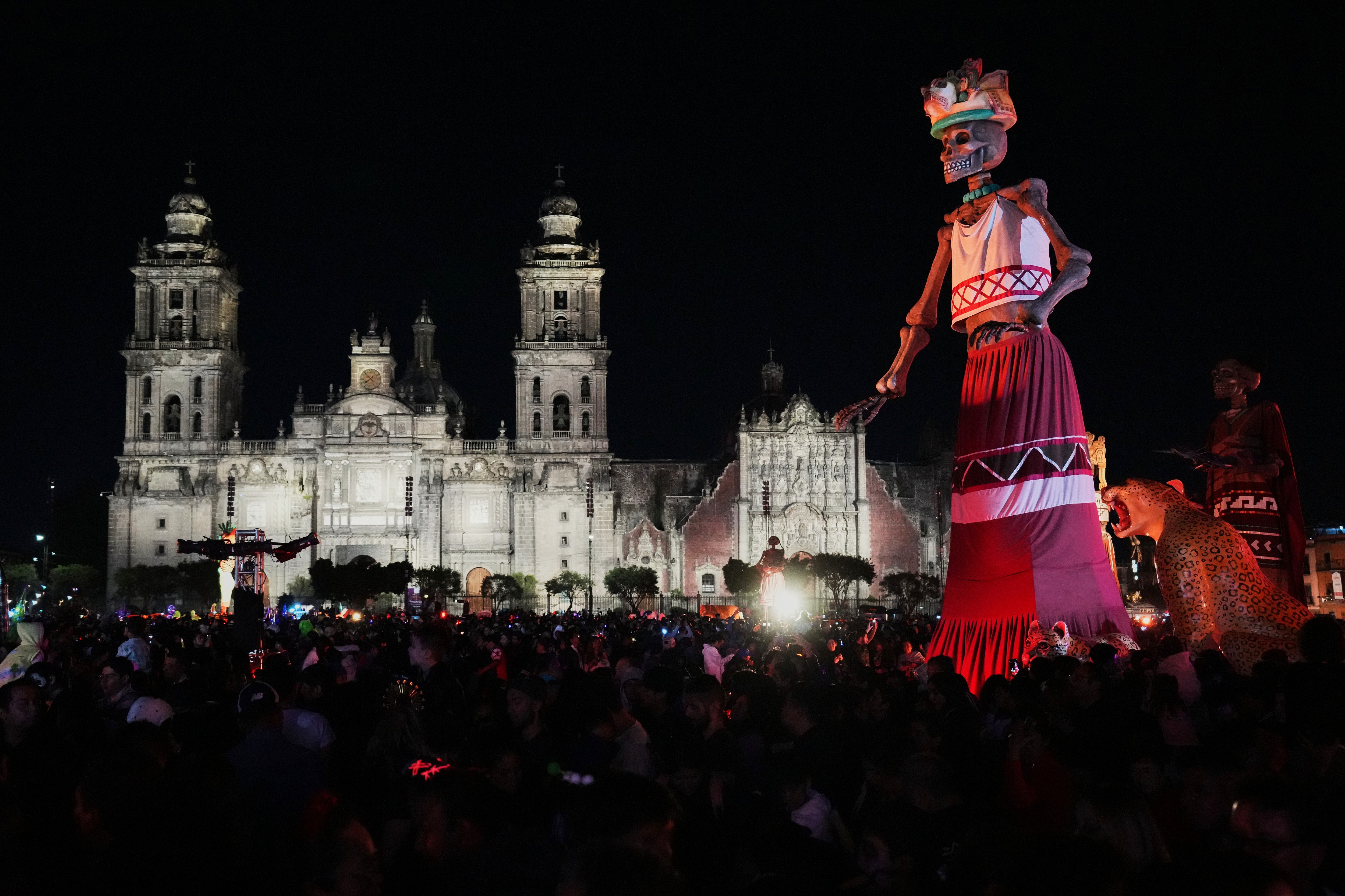 A crowd gathers around a very tall statue of a skeleton in a town square in Mexico City.