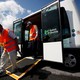 People in orange vests use a ramp to exit a driverless bus labeled "Robot Shuttle."
