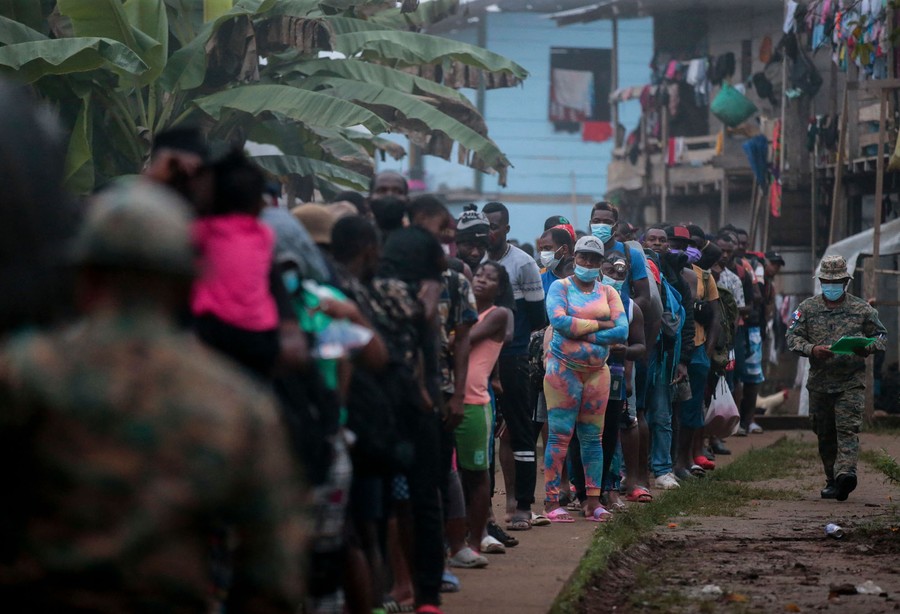 People stand in a long line in a vilage, waiting for transport.