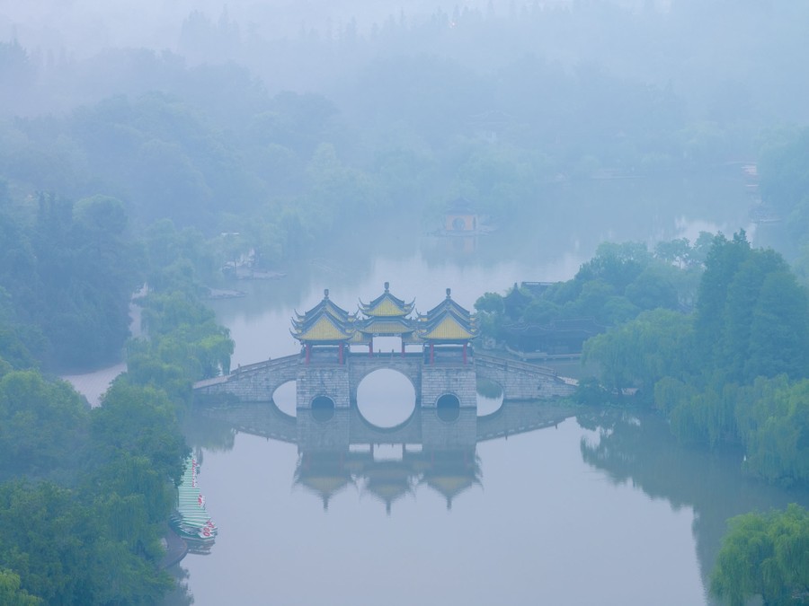 A historic covered bridge spans part of a lake in China, seen on a misty day, from above.