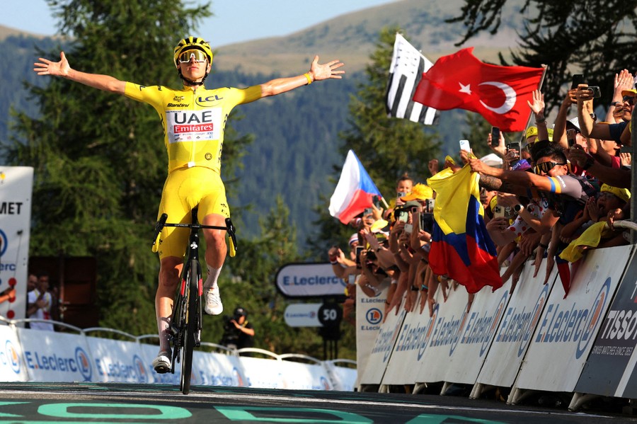 A cyclist in a yellow jersey holds his arms out as he crosses a finish line, cheered by a crowd on the sidelines.