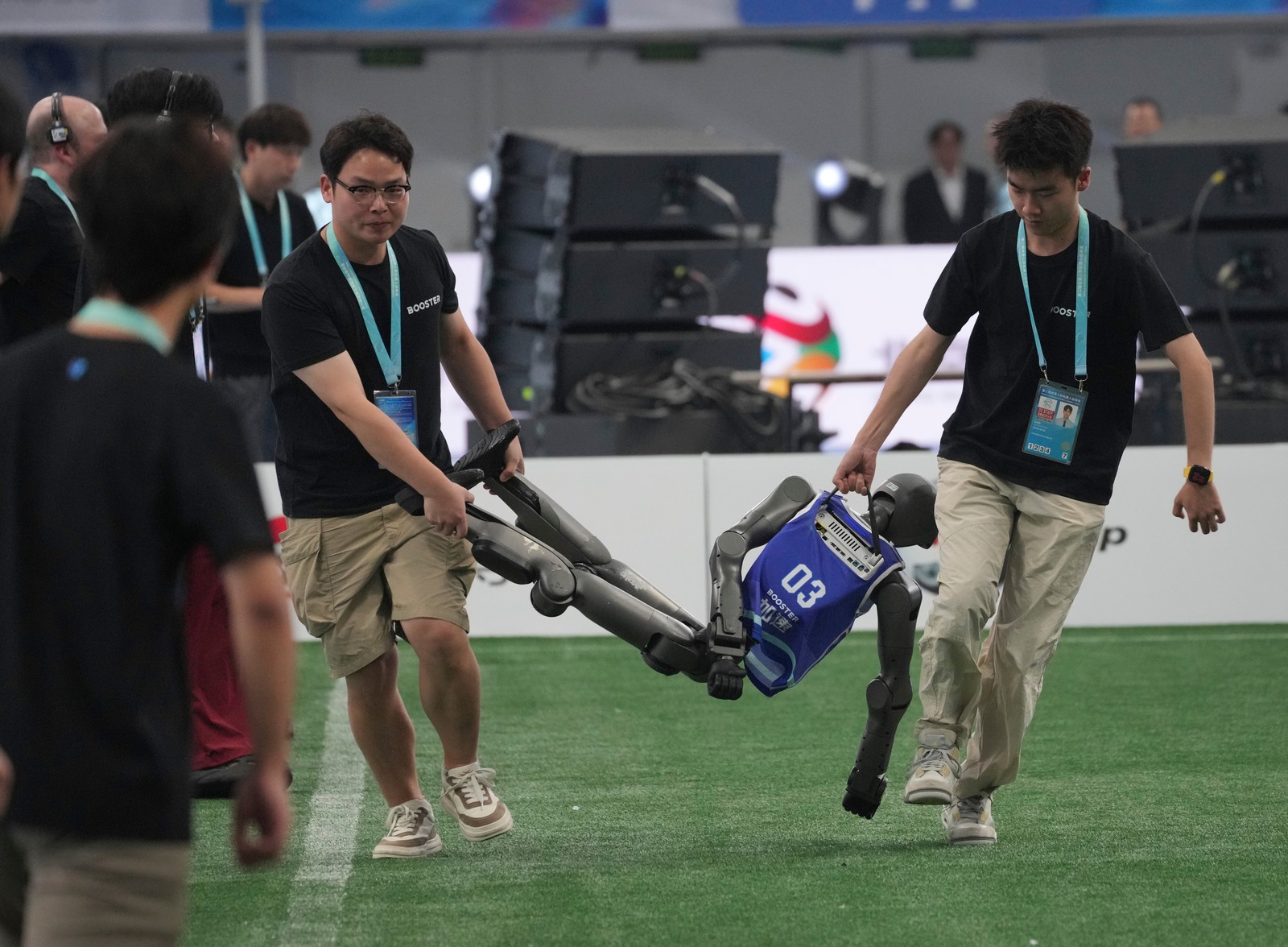 Two people carry an inert humanoid robot off a soccer field.