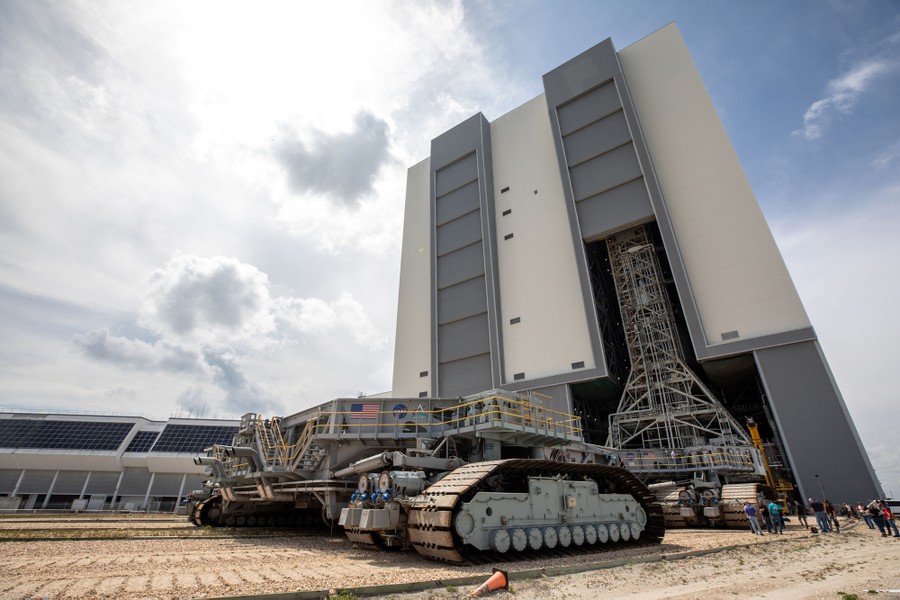 A gigantic tracked vehicle moves slowly toward a tall building to get underneath a rocket.