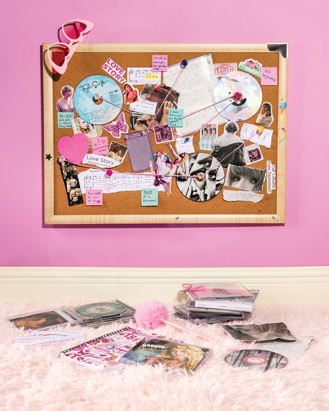 A cork bulletin board covered in Taylor Swift CDs, photos, and ephemera connected with pink string, with heart-shaped sunglasses hanging from one corner