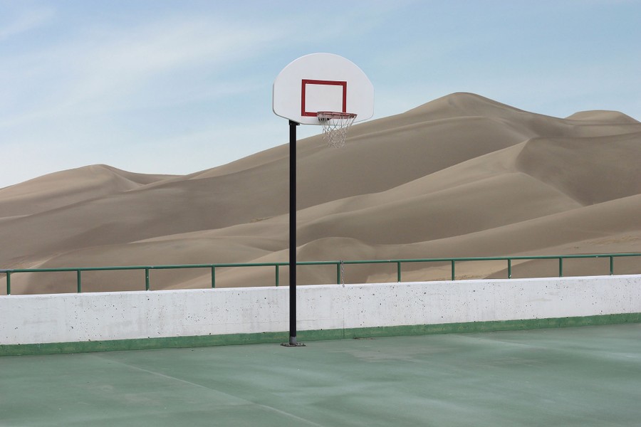 A basketball hoop stands in an empty court, seen before large sand dunes.