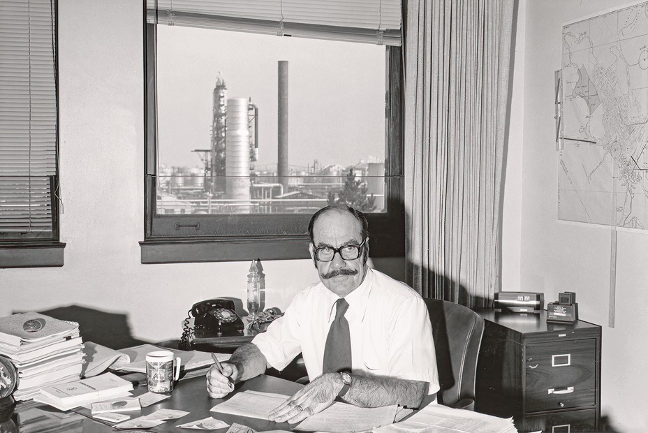 black and white photo of balding man with mustache and dark-framed glasses at desk holding pen with file cabinet, rotary-dial phone, and window in background looking out on a refiinery