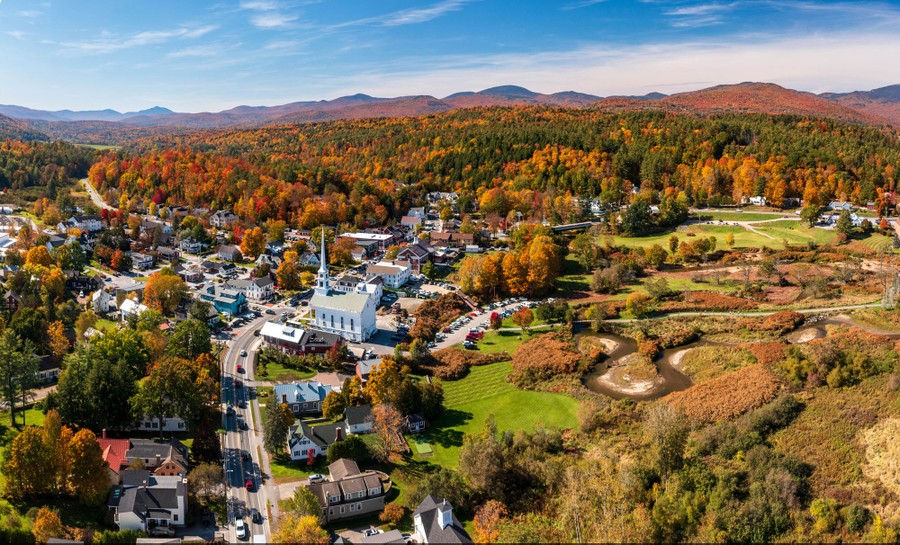 An aerial view of a town in the fall, featuring a white church steeple, surrounded by wooded mountains
