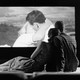 A black-and-white photo of a heterosexual couple watching another heterosexual couple kiss on a big screen