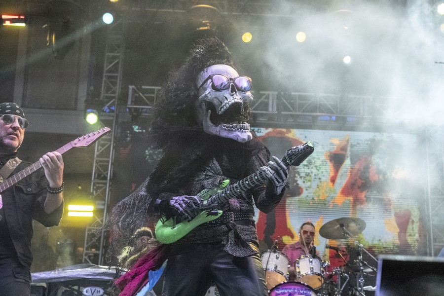 A person wearing a costume with a large skull mask plays guitar with a band on a stage.
