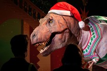 People look at a life-size T. rex sculpture dressed in a Santa hat and festive sweater.
