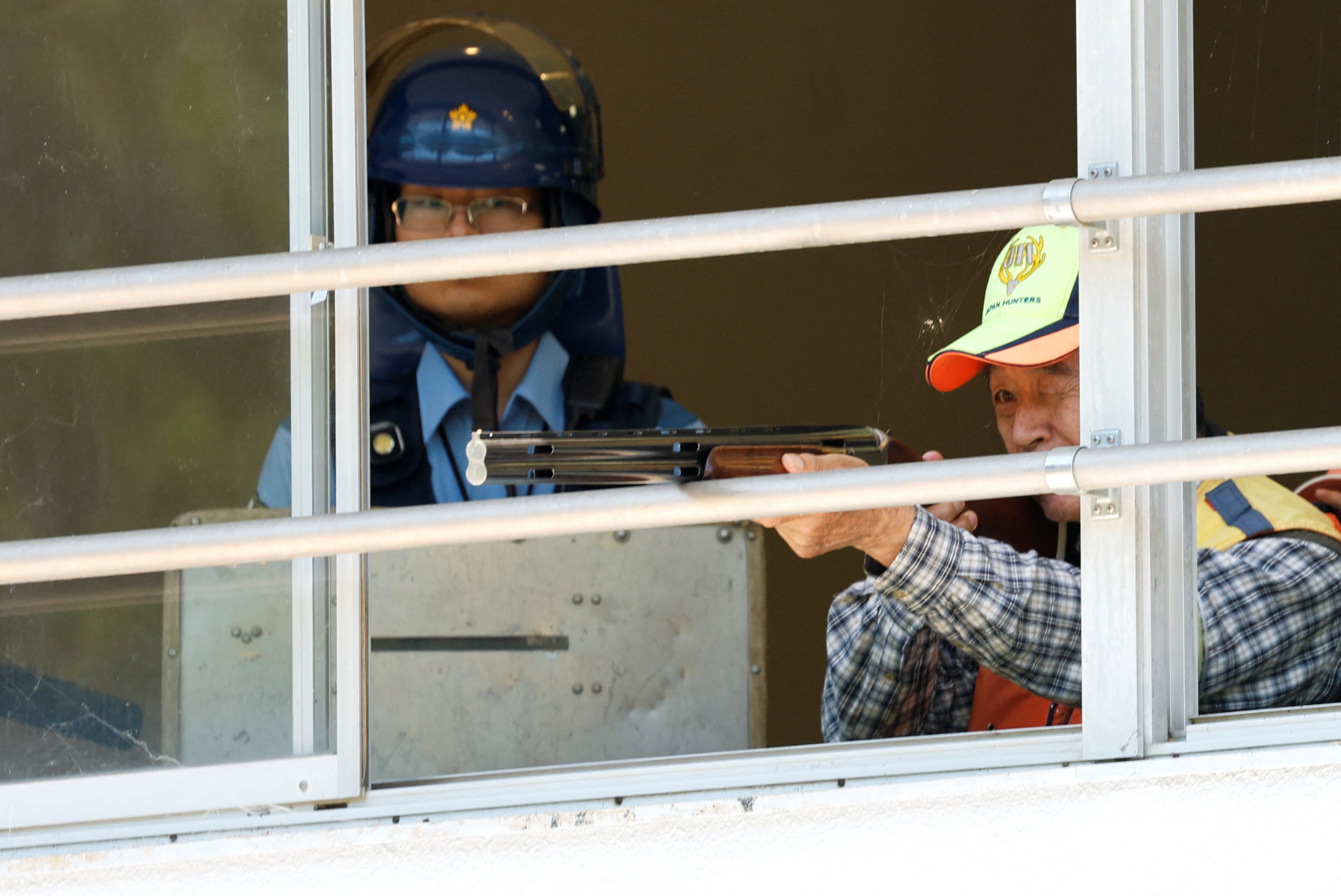 A hunter points a rifle through an open window, with a police officer watching nearby.