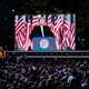A crowd stares up at President Donald Trump while he makes a speech behind a podium. Trump is visible on a giant screen to the crowd's left.