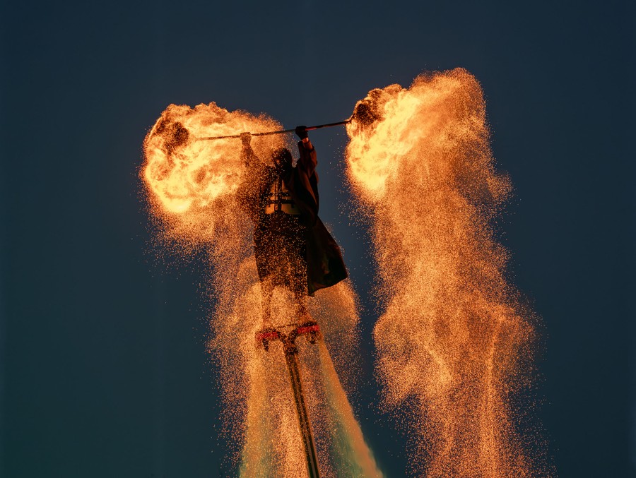 A performer hovers in the air, aboard a flyboard, holding a bar over their head with baskets of burning charcoal on either end, creating two streaming showers of sparks.