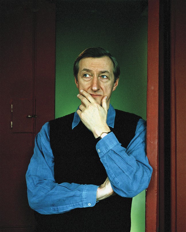 photo of man in blue collared shirt and black sweater vest standing in doorway with hand on chin