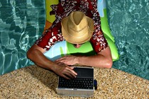 A photo of a man in a pool working on a laptop at the edge of the water.