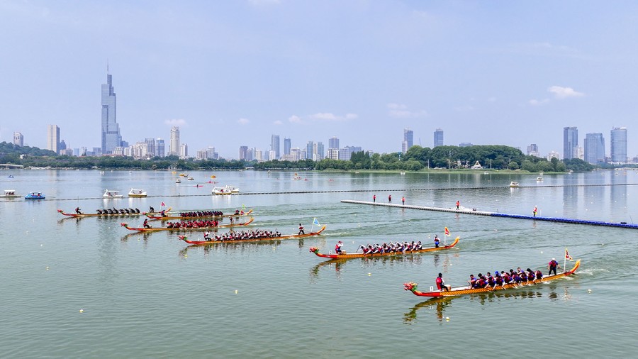 Six long dragon boats begin a race on a lake, with a city skyline in the background.