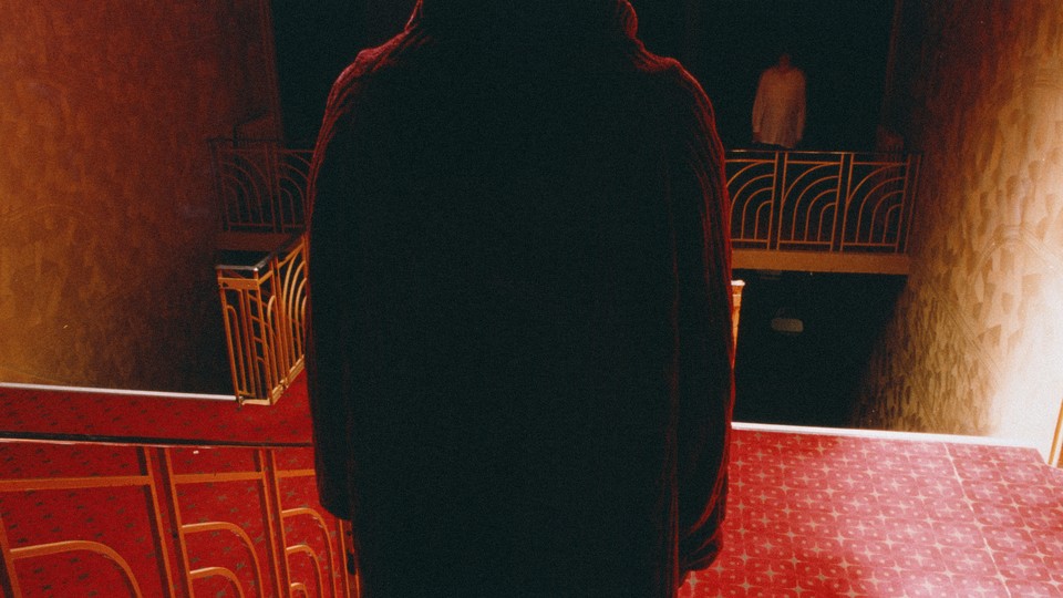 Filling the middle of the frame is the back of a person standing on a staircase, facing another person across the hall. The carpet is red, and shadows are deep.