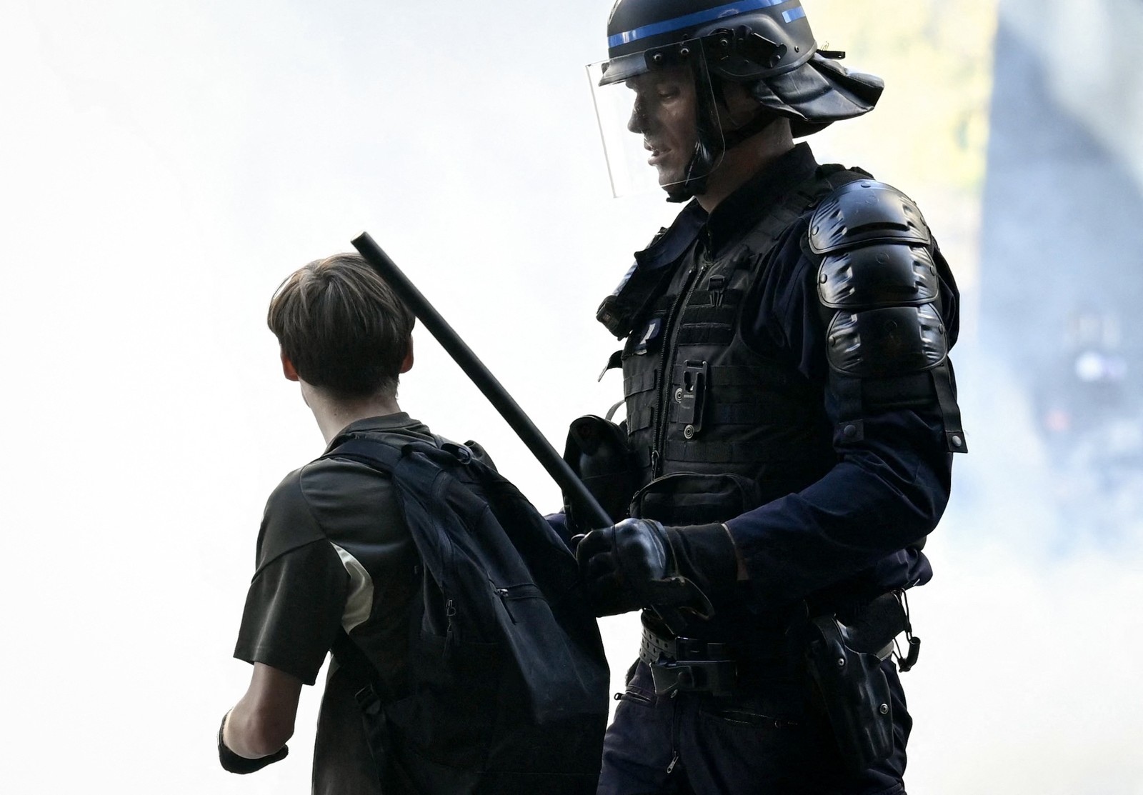 A riot police officer in full gear, holding a baton, stands much taller than a young person they are detaining.