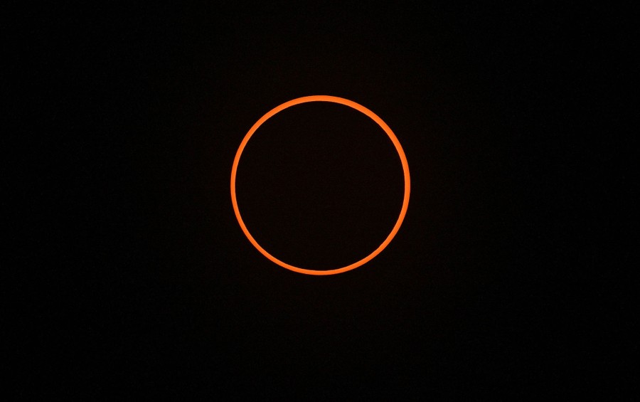 The moon appears in the sky directly in front of the sun, slightly smaller than the sun, forming a distinct orange ring in a dark sky.