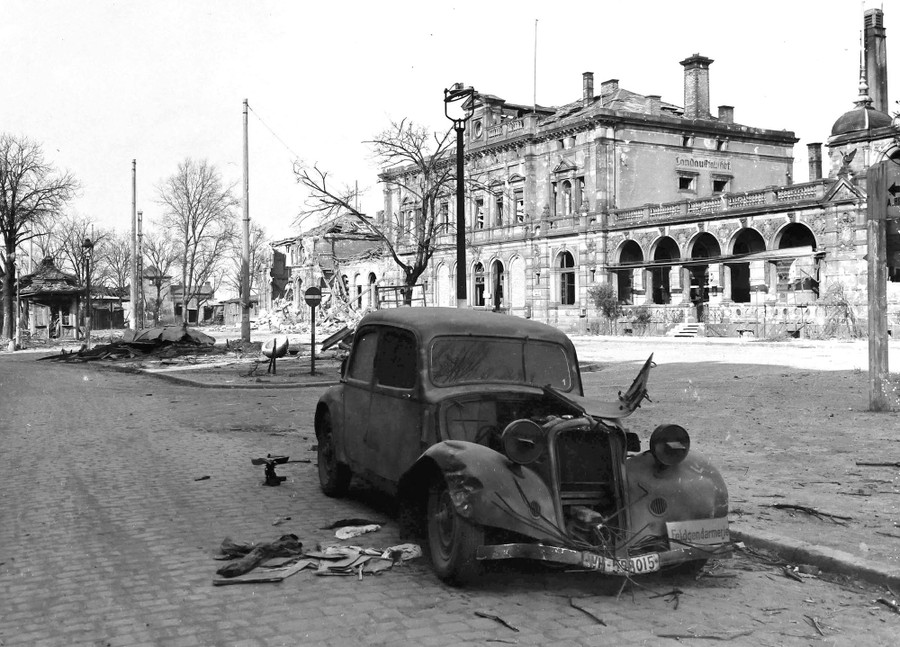 A smashed car sits in a road near a bomb-damaged railway station.
