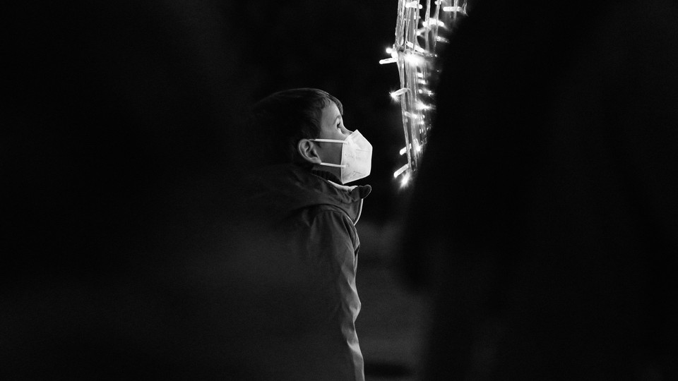 A little boy wearing a mask stares into glowing Christmas lights against a black background.