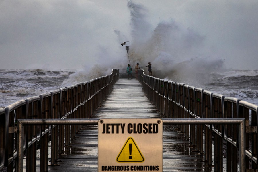 Three fisherman stand at the end of a jetty, surrounded by splashing water and churning waves. A sign at the entrance to the jetty reads "Jetty Closed: Dangerous Conditions."