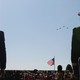 Photograph of two figures (left, Pete Hegseth) seen from behind, looking up at a fighter jet formation flying in a blue sky