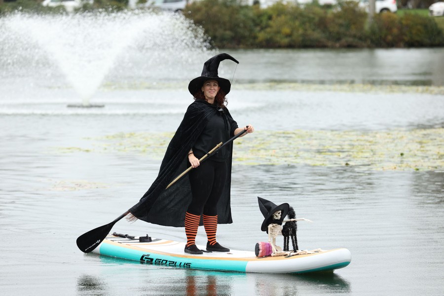 A person in a witch costume stands on a paddle board in a lake.